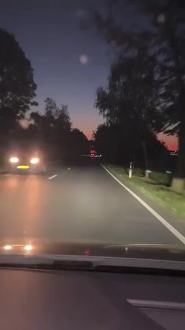 The video depicts a nighttime view from inside a vehicle driving on a paved road. The road is lined with trees on both sides, creating a tunnel-like effect. The car's headlights illuminate the road ahead, and there is another vehicle visible in the distance. A speed limit sign of 70 is visible on the right side of the road. The sky is transitioning from dusk to night, with a hint of orange still visible on the horizon.