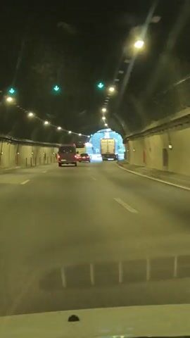 The video shows the interior of a tunnel from the perspective of a vehicle moving forward. The tunnel has a dark ceiling with rows of lights and green directional arrows. The walls are made of concrete and have vertical elements spaced out along them. There are several vehicles ahead on the road. Closest to the camera is a dark-colored van. Further ahead, there is a white panel van and a large white truck with a logo that appears to say "SALAMIS". The road is paved and appears to have multiple lanes. The tunnel opens up at the end, revealing daylight and trees outside. The video is short and captures the movement through the tunnel.