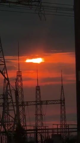 This video shows a sunset over a power substation. The sun is a bright orange disk partially obscured by clouds. The sky is a deep red and orange. In the foreground, there are several tall metal towers with electrical wires strung between them. Silhouetted against the sky are trees and some structures that appear to be buildings or vehicles. The video is shot from a low angle, looking up at the sky and the power substation.