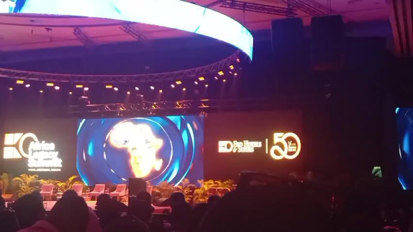 The video shows a large stage setup for a conference or event.  There are multiple large screens displaying logos and branding, including "African Legacy Summit" and "Radisson Hotels & Suites 50." The stage is lit with colorful lights, including a large circular lighting rig overhead. There are rows of chairs facing the stage, with people sitting in them. The video appears to be taken from the audience perspective. The overall environment suggests a professional and celebratory event.