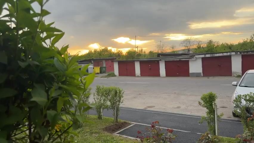 The video opens with a close-up view of green foliage, then slowly pulls back to reveal a static outdoor scene at sunset. A long row of detached garages with white walls and red doors stretches across the midground. In the foreground, there is a grassy area with small green bushes and a paved walkway/parking spot. A white Hyundai i20 car, bearing the license plate "CJ 09 DDK," is parked on the right side of the frame. To the far left, near the garages, three trash bins (yellow, green, and blue) are visible. The sky above is cloudy, with a bright orange glow of the setting sun breaking through. The background features more distant trees and what appears to be a residential or industrial area. The overall impression is a quiet, typical suburban environment at dusk, consistent with the provided context of Târgu Mureș, Romania.