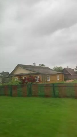 The video displays a series of houses and fences in a rural setting, possibly a village. The footage is shaky, suggesting it was filmed from a moving vehicle. The sky is overcast, indicating cloudy weather. The houses vary in architectural style and condition, with some appearing older and made of wood, while others are more modern with stone or brick facades. Fences of different materials, including wood and a patterned metal, separate the properties. Green grass and some trees are visible, contributing to the bucolic atmosphere.