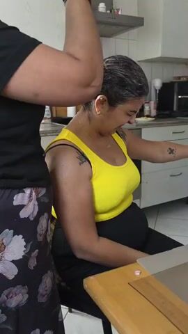 The video shows two women in a kitchen setting. One woman, wearing a black t-shirt and floral patterned pants, stands and applies hair dye to the other woman, who is sitting and wearing a yellow tank top and black pants. The woman applying the dye is wearing black gloves and holding a pink-capped bottle of hair dye. The seated woman is looking down and to her right with a slight smile, and has tattoos on her arm. The kitchen appears to be a typical home kitchen, with cabinets, a stove hood, and a table visible.