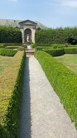 A gravel path winds through a manicured garden. On either side of the path are perfectly trimmed green hedges. In the distance, a classical-style alcove features a statue and a small fountain. A large, dark-colored building with a sloping roof is visible behind the hedges. The sky above is clear and blue, suggesting a sunny day.