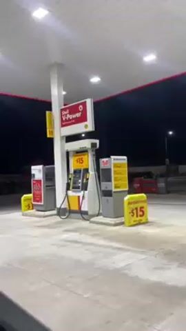 The video shows a Shell gas station at night. The station has a white roof with red trim, supported by a white column. Underneath the roof, there is a Shell V-Power sign and gas pumps. The pumps are white and yellow with "15" displayed on them. There are also gray payment kiosks on either side of the pumps. Yellow signs that say "Autoservicio $15" are placed around the pumps. The ground is gray concrete. The background is dark, with a few lights visible in the distance. A red truck is parked in the background to the left.