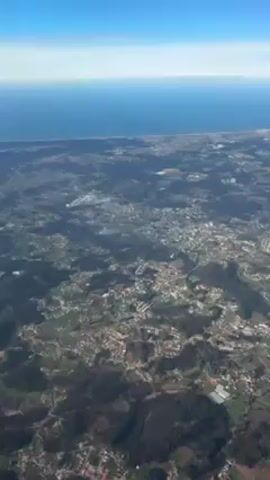 The video shows an aerial view of a coastal area, with the blue ocean stretching to the horizon under a clear, blue sky. A narrow strip of land marks the coastline, beyond which lies a landscape dotted with buildings, trees, and open spaces, appearing as a mix of urban and rural areas. The terrain seems slightly hilly or undulating, with clusters of vegetation scattered throughout the built-up environment.