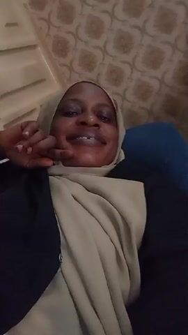 A woman with dark skin and wearing a light beige headscarf and a dark blazer is smiling at the camera. She is reclining in what appears to be a chair or couch. Her left hand is raised near her face. The background is a beige patterned wallpaper and a white object with lines. The image is taken from a low angle.