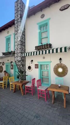 The video captures a sunny daytime scene along a charming, narrow street, likely in a town or village. The focus is on an outdoor cafe or restaurant with white-washed buildings, featuring vibrant turquoise window frames and doors, and a traditional red-tiled roof. An ornate black metal structure runs vertically up the side of the main building, adding a unique architectural detail.

Outdoor seating areas are set up on the multi-colored paved sidewalk, with numerous wooden tables and chairs in various hues including yellow, pink, and light blue, some with striped cushions. A green and white striped awning provides shade over a section of the tables.

Several individuals are present. One woman, dressed in a yellow top, is seated at a wooden table, seemingly engrossed in work on a laptop. Another person is seen walking away from the camera further down the street. Under a canopy of lush purple bougainvillea, another person is seated at a table.

The sky is clear and bright blue, with a strong sunbeam causing a lens flare. The light casts distinct shadows, indicating the middle of the day. Abundant purple bougainvillea blooms add a splash of color to the scene, draping over balconies and entrances. Green trees line the street in the distance. The overall impression is one of a peaceful, leisurely day in a picturesque Mediterranean setting. No discernible text is visible on any signs or buildings.