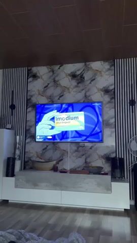 A television screen displays an advertisement for Imodium. The ad shows a hand holding a box of Imodium tablets and features animated characters. The background of the television is a marble-patterned wall with vertical decorative panels on either side. Below the television is a white media console with a grey textured top. A large wooden bowl sits on the console, along with other decorative items. The ceiling is paneled with wood. The overall environment appears to be a living room.
