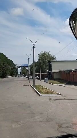 The video is a first-person perspective shot from inside a vehicle, likely a truck, as evidenced by the dashboard visible at the bottom of the frame and the large side mirror in the upper right corner. The vehicle is moving forward on an asphalt road. On the right side of the road, there is a fenced area with a brick wall. There are also streetlights and some buildings. On the left side of the road, there are trees and more buildings. The sky is blue with scattered clouds. There are a few cars and a large semi-trailer truck visible on the road. A trash bin is also visible on the right side of the road. The overall scene appears to be an urban or industrial area on a sunny day.