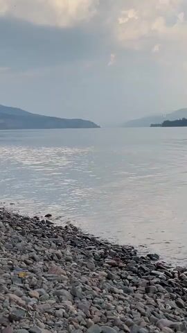 A slow pan shows a rocky shore leading to a wide, calm river. The river reflects the overcast sky and distant, hazy mountains that frame the horizon. The clouds are a mix of grays and whites. The water is still, with only slight ripples near the shore. The overall atmosphere is serene and tranquil.
