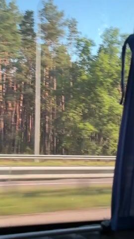 The video is a blurry, first-person perspective shot from inside a moving vehicle, likely a bus or car. The view is through a window showing a landscape of trees and a highway. The trees are predominantly green with some taller, thinner trunks visible, suggesting a forest. There is a concrete barrier and a roadway visible below the trees. The speed of the vehicle causes significant motion blur, making specific details difficult to discern. In the latter part of the clip, a small, light-colored structure with an arched opening, possibly a bus stop shelter, passes by on the right side of the road. The sky is visible and appears blue with some sunlight. The overall impression is of a scenic drive through a wooded area.