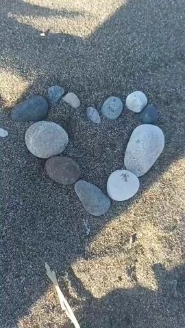 A close-up, overhead shot captures a heart shape formed from smooth, gray stones on dark, sandy ground. The stones vary in size and shape, with some larger and rounder, and others smaller and more elongated. Sunlight casts shadows across the scene, highlighting the texture of the sand and stones. The background is blurred, suggesting a shallow depth of field.