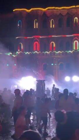 This is a vertical video of a nighttime outdoor event.  In the background, a multi-story building is illuminated with colorful lights arranged in arched patterns.  The foreground and midground are filled with a crowd of people of various ages, many of whom appear to be engaged in a playful water activity, as indicated by the mist and water spray visible.  The ground appears wet and reflective, suggesting it is a stage or a specially prepared area for the event.  The lighting is dramatic, with bright spotlights creating a hazy, atmospheric effect.