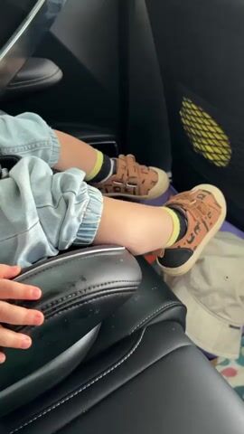 The video shows a close-up view of a child's legs and feet inside a vehicle. The child is wearing light blue jeans, yellow and black striped socks, and brown shoes with a tiger pattern. The shoes have velcro straps. The child's legs are resting on a black car seat. In the background, a mesh pocket with a yellow object inside is visible on the car door, and a white hat and a blanket are on the floor. The lighting suggests an indoor environment, likely the interior of a car. The video is static, with no movement or dialogue.