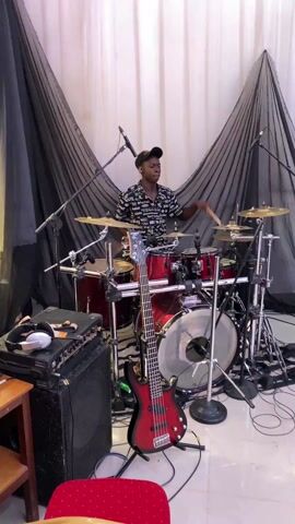 A young man plays a drum set indoors. He wears a black baseball cap and a black and white patterned button-up shirt. He hits the drums with drumsticks, and looks focused on his playing. The drums are red, and the set includes cymbals. A red bass guitar stands near the drums, and a black amplifier sits on the floor nearby. Microphones are positioned near the drums. The backdrop is white fabric with black fabric draped over it in two triangular shapes. The floor is white tile.