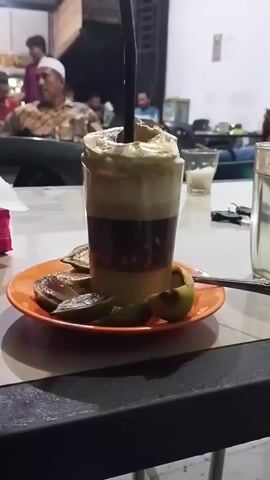 This is a video capturing a close-up of a layered drink in a clear glass, topped with a frothy cream and a sprinkle of cinnamon or cocoa powder. The drink is served on an orange saucer with several slices of lime or a similar citrus fruit. A dark straw is inserted into the drink.

In the background, the scene appears to be a casual outdoor or semi-outdoor establishment, possibly a cafe or a roadside eatery. The lighting suggests it's evening or nighttime. Several people are visible in the background, some seated and others standing. One man in the foreground, to the left of the drink, is wearing a white skullcap and a patterned shirt. He appears to be looking towards the camera or something beyond it. Other individuals are less clearly discernible but suggest a social gathering or a place where people are relaxing.

The setting has a rustic feel with visible structures and furniture typical of a local establishment. The overall atmosphere seems relaxed and informal. There is no visible text in the video. The video provides a close-up view of the beverage, highlighting its presentation, with a broader view of the surrounding social environment.