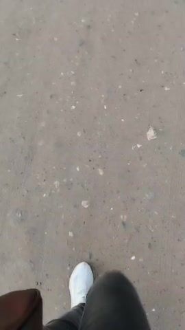 The video shows a low-angle, first-person perspective of someone walking on a paved surface, possibly a sidewalk or road. The camera is pointed downwards, focusing on the lower legs and feet of the person walking. The person is wearing white sneakers and dark-colored pants, possibly leggings or jeans. A brown bag or backpack is visible on the left side of the frame, hanging from the person's side. The ground is a neutral, sandy or concrete color with small pebbles and debris scattered on it. The footage is slightly blurred, indicating movement. The timestamp indicates the video is very short, only a few seconds long.