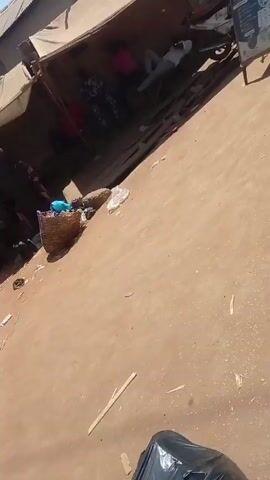 The video shows a man walking by a roadside stand. The man is wearing jeans, a grey shirt, and sandals, and he is carrying a box and another object. He walks from right to left in the frame. The roadside stand has a sign that reads "EXCEL COMMUNICATION GET YOUR IOS MACHINES HERE." There are people sitting and lying down near the stand. A motorcycle is parked there as well. The area appears to be a somewhat crowded street or marketplace. A loud horn sounds during the video.