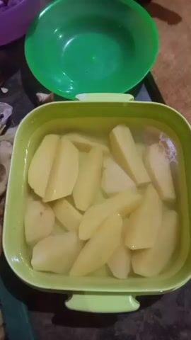 The video shows a close-up, top-down view of a light green plastic container filled with water and potato wedges. The potatoes are cut into wedges, some are whole, and others are cut in half. There is a green plastic bowl in the background. The scene appears to be in a kitchen or food preparation area. A hand is visible on the right side of the frame, resting on the container. The overall impression is one of food preparation, with the potatoes likely being soaked or washed.