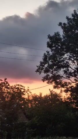 The video displays a time lapse of a sunset over a rural landscape. In the foreground, a white house with a pitched roof and dark windows is partially visible on the left. Trees with dark foliage frame the right side of the scene, with some trees in the middle ground. Overhead, power lines stretch horizontally across the frame. The sky is filled with dramatic clouds, transitioning from dark grey at the top to vibrant hues of orange and yellow near the horizon, indicating the setting sun. The overall atmosphere is serene and picturesque.