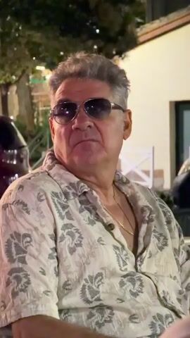 A man with graying hair and sunglasses is sitting outside. He is wearing a patterned button-down shirt and a gold chain. The background shows an outdoor seating area with trees and buildings.