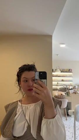 A young woman is taking a selfie in a mirror. She has dark, curly hair pulled back and sunglasses perched on top of her head. She is wearing a white puffed-sleeve blouse with a brown and white checkered vest over it. A silver chain is visible around her neck. In her right hand, she holds a smartphone, which she is using to take the photo. Her left hand is holding a pair of pink headphones.

The setting appears to be a brightly lit indoor space, possibly a salon or a shop, with a clean, modern aesthetic. In the background, shelves are filled with rows of colorful bottles, likely nail polish. There is a small round wooden table and a white fan visible. The flooring is a speckled pattern. The overall mood is casual and perhaps a bit playful, as indicated by her smile. The lighting suggests it is daytime. The location context of Suthep, Thailand, provides a geographic anchor to the scene.