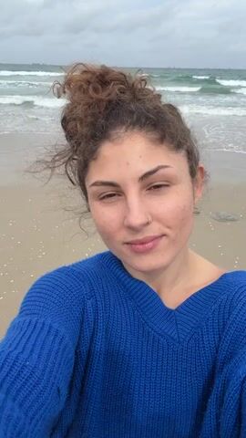 The video shows a woman with curly brown hair in a messy bun, wearing a blue sweater, standing on a sandy beach. She has fair skin, brown eyes, and a nose ring. She smiles and looks directly at the camera. Behind her, the ocean waves crash onto the shore under a cloudy sky. In the distance, ships can be seen on the horizon. The sand is wet, and small white shells are visible. The wind is blowing, causing some of her hair to fly around her face. The overall impression is that the woman is taking a selfie at the beach on a cloudy day.
