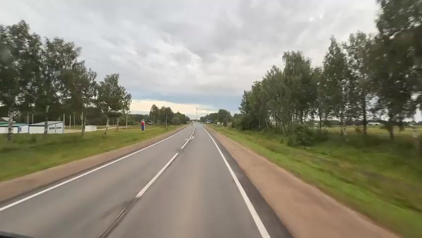 The video is a first-person perspective driving through a rural landscape under a cloudy sky. The road is paved with a white line marking the lanes. Trees line both sides of the road. There are a few buildings and signs visible. A few cars and a truck are seen on the road. One sign indicates a distance of 2.1 km to "ST. OSTROVSKOE".