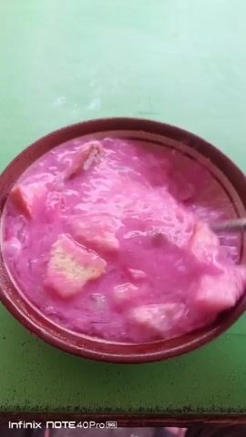 A close-up overhead shot shows a pink-colored, creamy dessert in a brown bowl. The dessert contains pieces of what appear to be bread or pastry, along with possibly some fruit or other solid ingredients. A spoon is partially submerged in the dessert, lifting a portion of it, indicating that it is being served or eaten. The bowl is placed on a green, textured surface. The bottom of the frame displays a watermark that reads "Infinix NOTE 40 Pro". The lighting suggests an indoor or shaded outdoor setting.