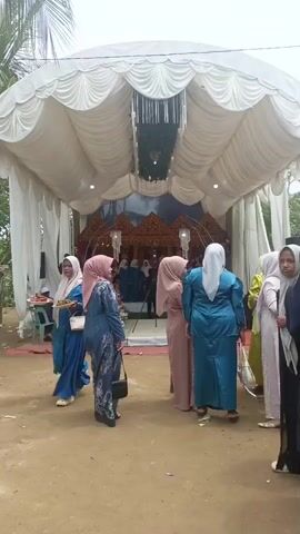 The video shows an outdoor event, likely a wedding reception, under a large white tent. Several women in hijabs and traditional clothing are standing and mingling in front of the tent. One woman on the left is holding a tray of food. Inside the tent, there appears to be a decorated stage with people sitting. The ground is dirt, and there are some trees visible in the background. The overall atmosphere seems festive and social.