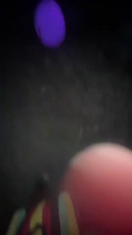 The video shows a dark outdoor scene with various colored lights illuminating the area. There are several glowing orbs of different colors, including purple, pink, and green, scattered throughout the scene. Some of these orbs are suspended from trees or branches, while others are on the ground. The ground appears to be covered in leaves and debris. The overall impression is that of an art installation or a decorated outdoor space at night.