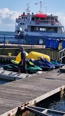 A man in a wetsuit stands on a floating dock next to a jet ski. He is wearing a cap and appears to be adjusting his goggles or mask. Behind him, two large yellow buoys are visible, and beyond that, a large white boat named "AMBER EXPRESS" is docked. The water is choppy, and the sky is partly cloudy.