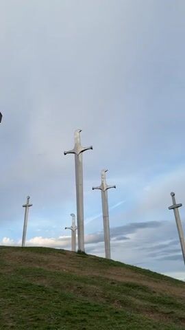 The video shows a grassy hill with several large metal sculptures of swords sticking out of the ground. There is also a large stone structure resembling a cross or "T" shape. A group of people is visible at the top of the hill near the stone structure, and one person walks down the hill towards the camera. The sky is cloudy with patches of blue.