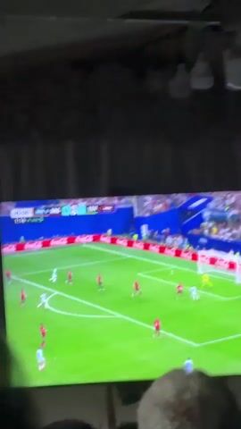 A large television screen displays a soccer match, with two teams in red and white uniforms on a green field. The score indicates Argentina leading Egypt 1-0. In the foreground, several people are seated, facing the television. The camera then pulls back to reveal a large gathering of people seated in a tent-like structure, also watching the soccer match on the television. The environment appears to be an outdoor or semi-outdoor venue, possibly a covered patio or a large tent, with fabric draped from the ceiling and lights hanging down. The overall scene depicts a group of people collectively watching a sporting event.