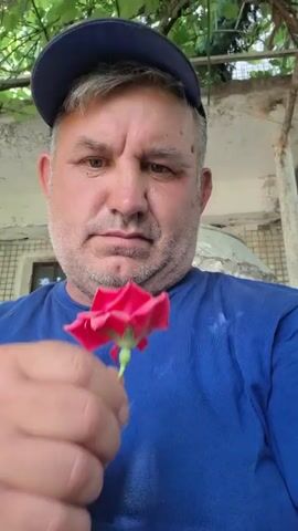 A middle-aged man wearing a blue t-shirt and a dark blue baseball cap holds a single red rose near his face, bringing it to his nose as if to smell it. The background is out of focus but appears to be outdoors, with green foliage visible above the man's head and a building with a tiled wall behind him. The man's expression is serious and his gaze is directed towards the camera. The rose is held between his thumb and forefinger, with the bottom of the flower, including the stem and sepals, facing the camera. The video then zooms in slightly on the rose.