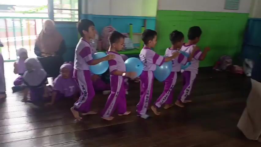 The video shows a group of young children in a classroom setting. The children are mostly wearing matching purple and white uniforms. Some children are seated on the floor, while others are standing and participating in an activity that involves balloons. The children are actively moving and interacting with each other, suggesting a playful or educational game. The room appears to be a typical classroom with wooden floors, tables, chairs, and backpacks. The overall atmosphere seems energetic and cheerful.