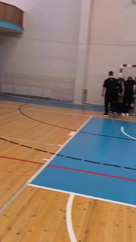 The video captures scenes inside an indoor gymnasium in Shaberdino, Russia. The gym features a wooden floor with visible red, white, and black court markings, including a basketball court layout. Blue-painted areas are present on the floor, with a center circle displaying the Cyrillic word "КУПОН" (Kupon).

Various groups of people, predominantly young adults and teenagers, are seen standing, walking, and conversing casually on the court. Some individuals are near a set of blue wall bars along one wall. Traffic cones are placed in a line on one side of the court, hinting at a structured activity or a setup for a competition.

In the background, there are basketball hoops and large banners with Cyrillic text. One banner prominently features "КУПОН" and other text, while another bears "НА СОРЕВНОВАНИЙ" (meaning "at competitions" or "for competitions"). A whiteboard is also visible. The atmosphere appears relaxed, with individuals engaged in conversations. The overall scene suggests a gathering, event, or the initial stages of a competition in a school or community sports hall.
