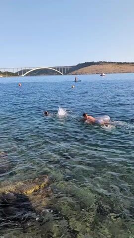 Two people are swimming in the ocean near a rocky shore. One person is using a float and the other is swimming alongside. In the background, a bridge spans across the water, and there are several buoys and people on paddleboards. The sky is clear and blue.