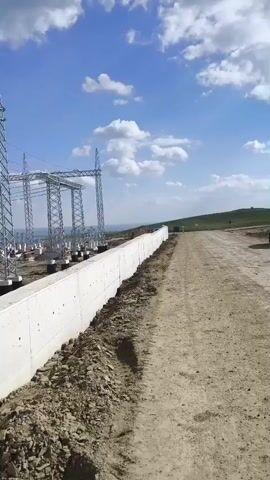 The video shows the construction of a power substation. Several tall metal transmission towers stand over concrete foundations. Some foundations are still under construction, with wooden forms visible. There is a long concrete wall running along the side of the construction site. The ground is mostly dirt and gravel. The sky is blue with scattered white clouds.