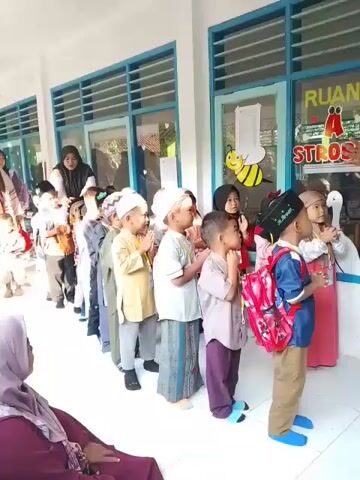 A group of children, mainly boys, are lined up single file along the corridor of what appears to be a school building. Most of the boys are wearing white religious caps. Some of the children have their hands together in a prayer-like gesture, while others are clapping. Several adults, presumably teachers or caregivers, are present. One woman is seated watching the children. The building has large windows with blue frames. A sign on one of the windows reads "RUANG STROBERI," which translates to "STRAWBERRY ROOM." There's a drawing of a bee on the window as well. The atmosphere seems to be one of anticipation or celebration.