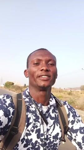 The video depicts a man, possibly in his late twenties or early thirties, facing the camera. He is wearing a blue and white floral-patterned shirt over a white undershirt, and a brown backpack. He has a short beard and dark hair. The background reveals a paved road, and arid landscape with scattered vegetation and what appears to be a power line structure in the distance. The sky is clear and bright, suggesting daylight. The man appears to be moving, possibly on a vehicle, as evidenced by the wind. He makes a puckered face, as if to blow a kiss.