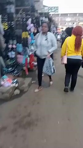 The video shows a busy outdoor market area. People are walking through the market, browsing the various stalls. The stalls are filled with colorful items, including shoes and clothing. The ground appears wet. The sky is overcast.
