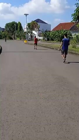 The video shows an asphalt road on a sunny day, with a few people walking on it. The sky is blue with scattered white clouds. Two people are visible closer to the camera, one wearing a blue shirt and shorts, and the other a red shirt. They are walking in the same direction, towards the right side of the frame. Further down the road, there are two more people walking in the same direction but are more distant. Houses and trees line the sides of the road. A solar-powered street light is visible on one side. A yellow object, possibly a trash can, is placed on the side of the road.