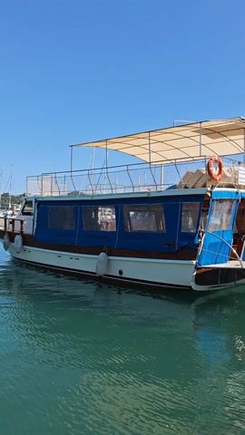 This video captures a bright, sunny day at a marina in Demre, Turkey. The scene is dominated by clear blue skies and calm, turquoise waters.

The video opens with a wide shot of a harbor filled with various boats, many of which appear to be tourist vessels or gulets, with distant hills forming the backdrop. A large boat with a white hull, wooden accents, and a blue canvas enclosure is prominent in the foreground.

The camera then offers a closer look at this particular boat, showing its beige sun canopy and a lifebuoy on deck. Following this, a group of people is seen walking along a wooden pier, heading towards a line of docked boats. Many of these boats, similar in design, are adorned with Turkish flags fluttering in the breeze.

The video then focuses on the details of these boats, showcasing their beige canopies, dark canvas enclosures, and safety equipment like lifebuoys. One boat prominently displays text in Turkish on its upper deck, indicating safety information: "TEKNENİN CAN YOLCU SAYISI: 40" (Maximum passenger capacity of the boat: 40), "CAN YELEĞİ SAYISI: 40" (Number of life vests: 40), and "CAN SİMİDİ SAYISI: 3" (Number of lifebuoys: 3). The Turkish flag is also visible on this boat.

The overall impression is one of a vibrant, active harbor on a beautiful day, likely bustling with tourists preparing for boat excursions.