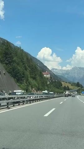The video is a first-person perspective shot from a vehicle traveling on a highway. The highway is paved and has multiple lanes, marked with white lines. The vehicle is in the right lane. On the left side of the highway, there is a steep, tree-covered mountainside. A chain-link fence runs along the edge of the highway, separating it from the embankment. Several vehicles, including cars and recreational vehicles, are parked or stopped on the shoulder or in a traffic jam on the left side of the highway. Further up the mountainside, a building with a red roof and a white facade, possibly a church or a small house, is visible. Beyond the immediate mountains, larger, snow-capped mountains can be seen in the distance. The sky is blue with scattered white clouds. The overall environment appears to be a mountainous region on a sunny day, with some traffic congestion.