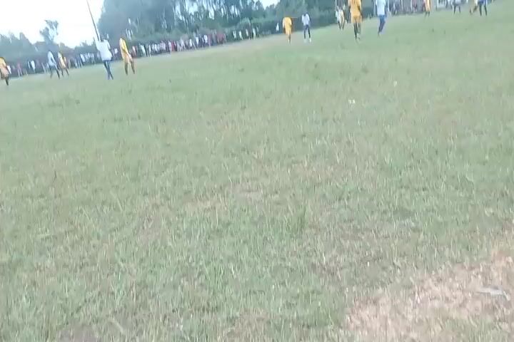 A soccer game is in progress on a grass field. Several players are visible, some wearing yellow shirts and others wearing white and blue shirts. A group of spectators is standing along the sidelines. In the background, there is a building, trees, and a radio tower. The sky is cloudy.