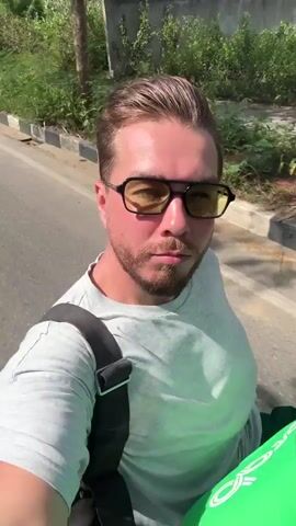 The video shows a man riding on the back of a motorcycle. The man is wearing a light gray t-shirt, black glasses, and a black shoulder bag. He has a beard and his hair is styled back. The driver of the motorcycle is wearing a bright green jacket with "Grab" printed on it. The background shows a road with greenery on either side, and a mountain range in the distance. The video appears to be shot from a first-person perspective by the man on the back of the motorcycle. The camera moves around as the motorcycle travels down the road. The audio includes the sound of the wind.