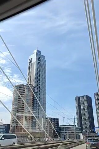 The video shows a view from a vehicle driving on a bridge with modern skyscrapers in the background. The sky is blue with some clouds. There are multiple lanes of traffic on the bridge, with cars visible. In the distance, there are several tall buildings, including a very tall, slender skyscraper and two darker, wider skyscrapers. There are also overhead wires, likely for an electric transportation system, and streetlights. The bridge has railings and a railing structure with cables. A person is briefly visible walking on the sidewalk or a platform below the bridge. The vehicle appears to be moving at a moderate speed.