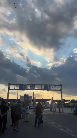 The video captures a wide shot of a border crossing with a dramatic sky filled with dark clouds and rays of sunlight. In the foreground, a group of people stand on a road, facing away from the camera towards a border checkpoint. The checkpoint has overhead signs with symbols indicating different types of vehicles and what appear to be passport control booths. A red traffic light is visible. In the background, several vehicles are lined up at the crossing, and there are structures that resemble gas stations. The overall impression is one of a busy transit point under a powerful sky.